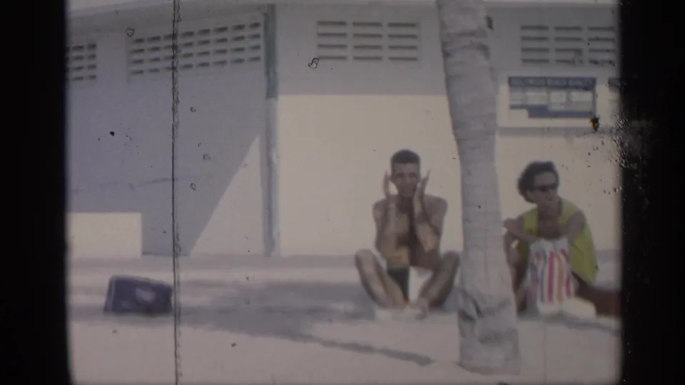 Two people in swimwear sit in shade near a palm tree and beach building in 1959 California home-movie footage.