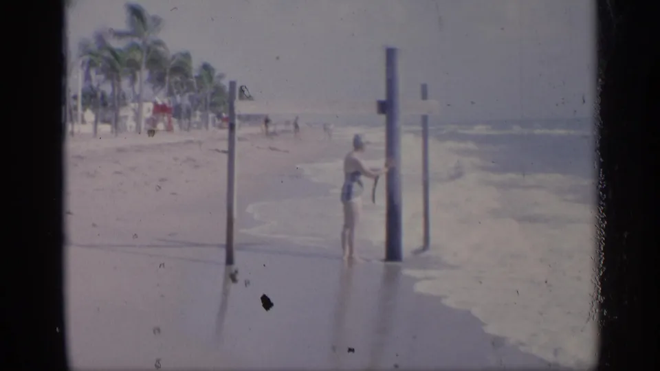 A swimmer stands beside shoreline posts as waves break nearby, with a palm-lined California beach in the background.