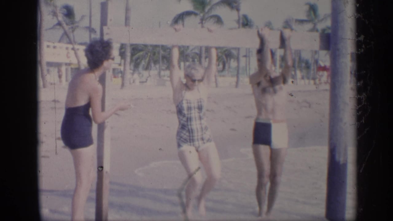 California 1959 home-movie frame showing two women watching as a man reaches up near a beach bar on poles.