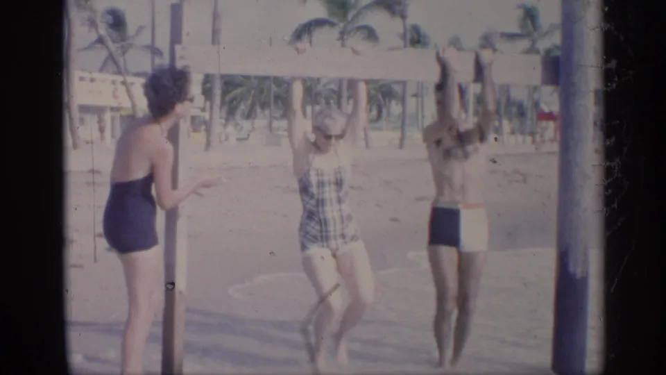California 1959 home-movie frame showing two women watching as a man reaches up near a beach bar on poles.