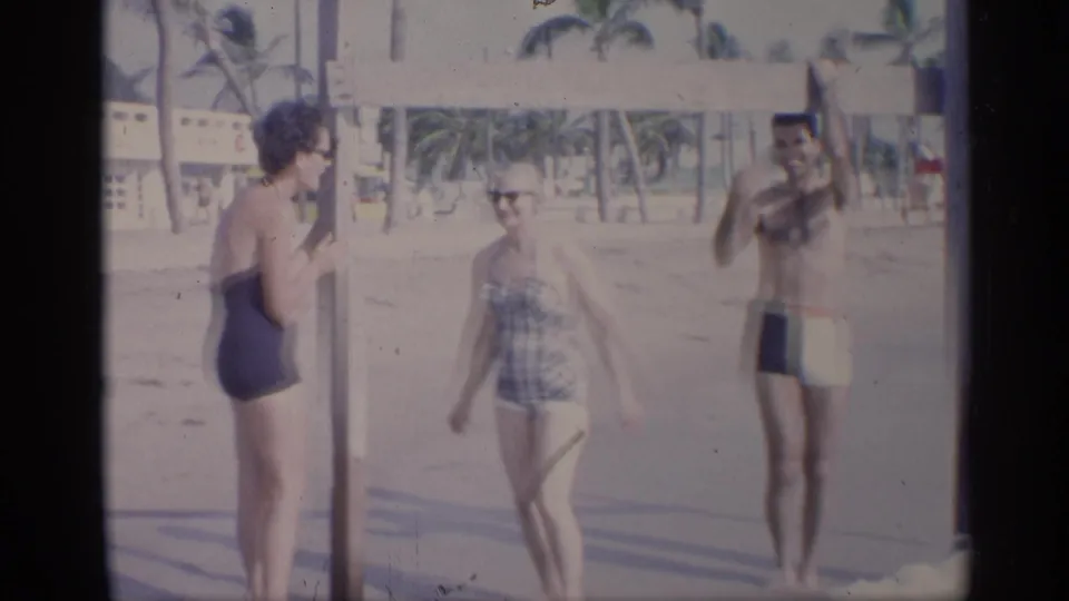 California 1959 beach scene with two women in one-piece swimsuits and a man in trunks near a pull-up bar.