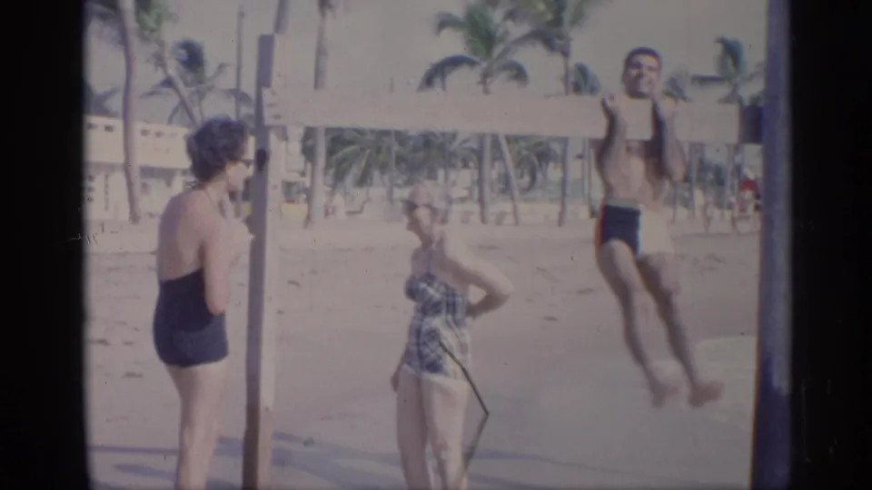 California 1959 frame showing a man mid-pull-up or jump at a wooden beach bar as two women stand nearby.