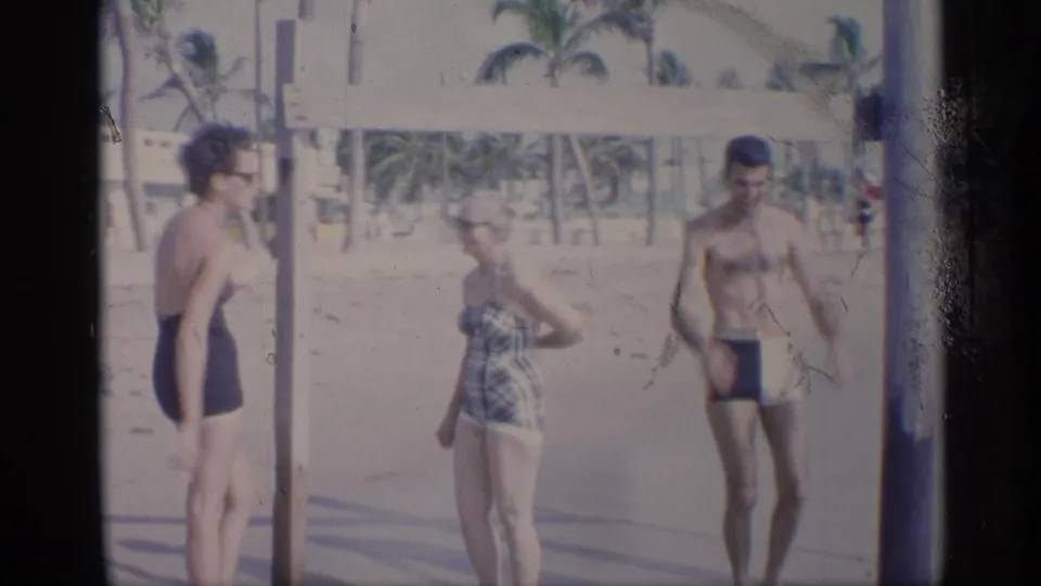 California 1959 home-movie frame with three adults beside a beach exercise bar, one woman turning toward the man.