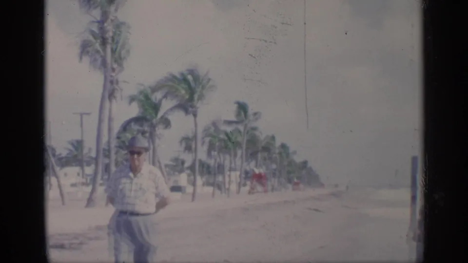 1959 California beach home-movie frame with a man in a light shirt and hat near palms, sand, and a hazy shoreline.