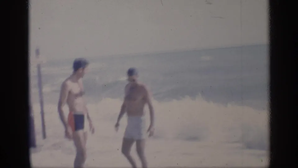 1959 California beach home-movie frame with two men in swim trunks walking along the surf line beside posts and waves.