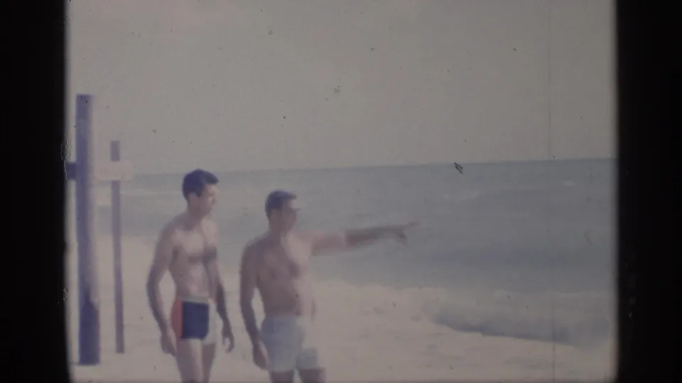 1959 California beach frame with two men standing near waves, one gesturing toward the water as the surf rolls in.