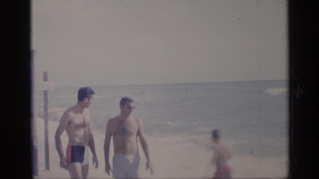 Two shirtless men in swim trunks walk along a beach as surf rolls behind them.