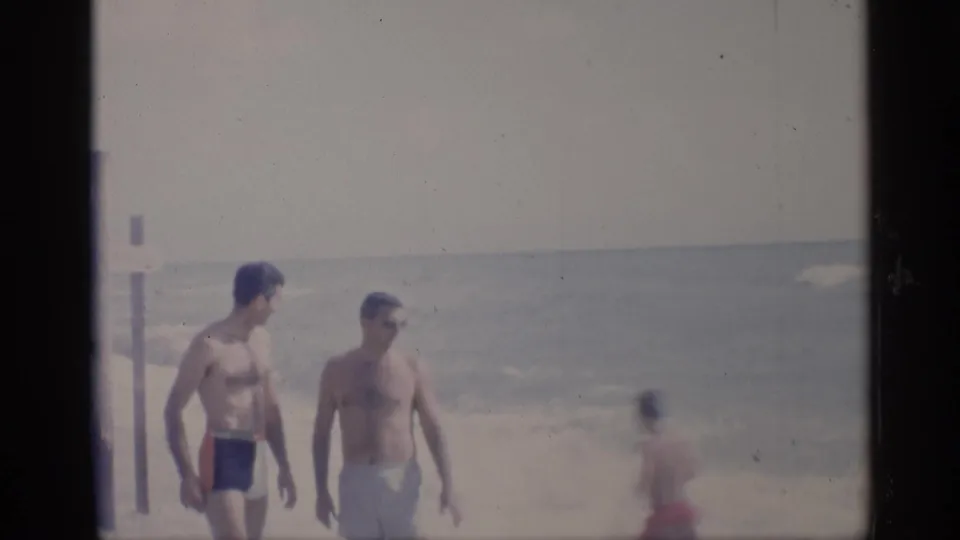 Two adult men in vintage swim trunks walk on a California shoreline, with ocean waves and a beach post visible behind them.