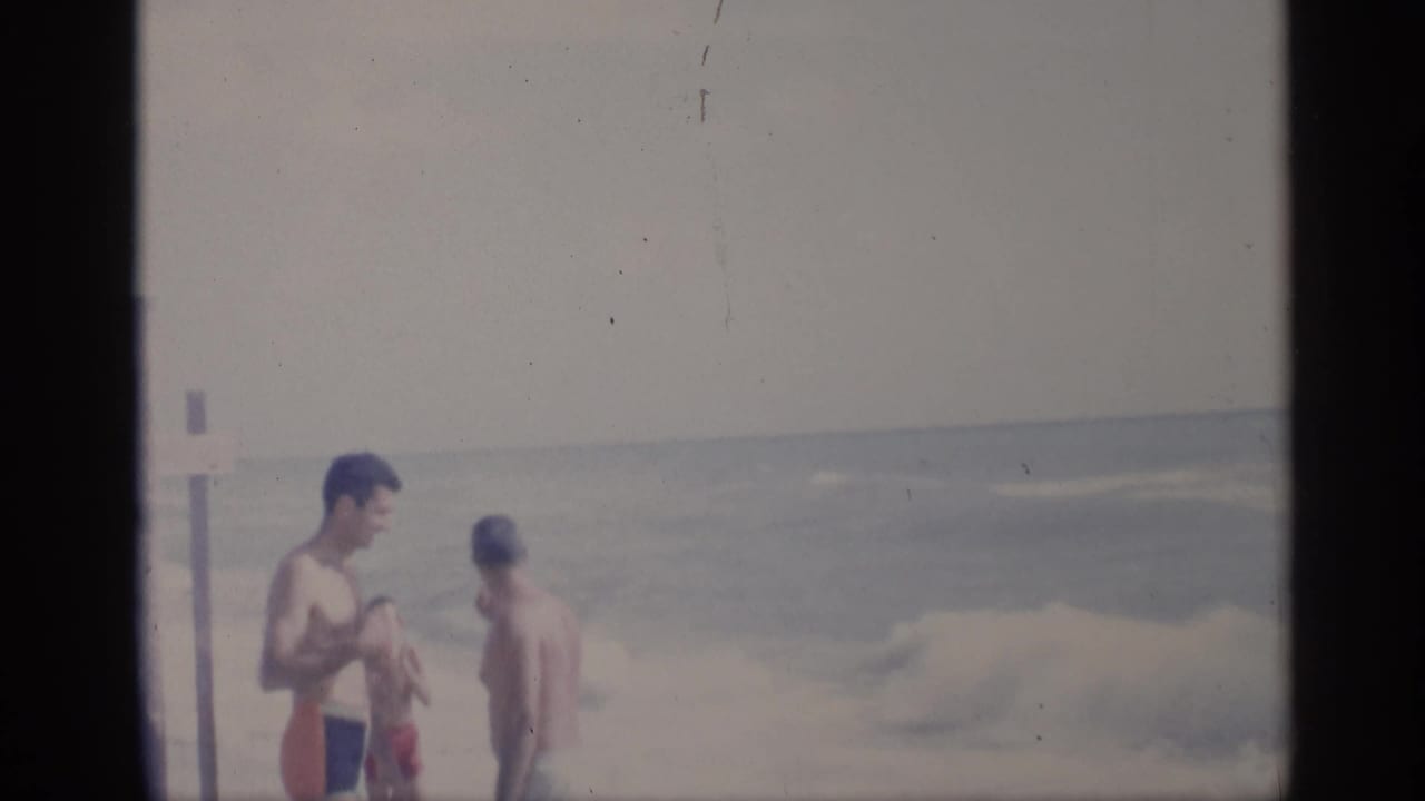 Two men stand near the water while a child-sized blurred figure passes in front.