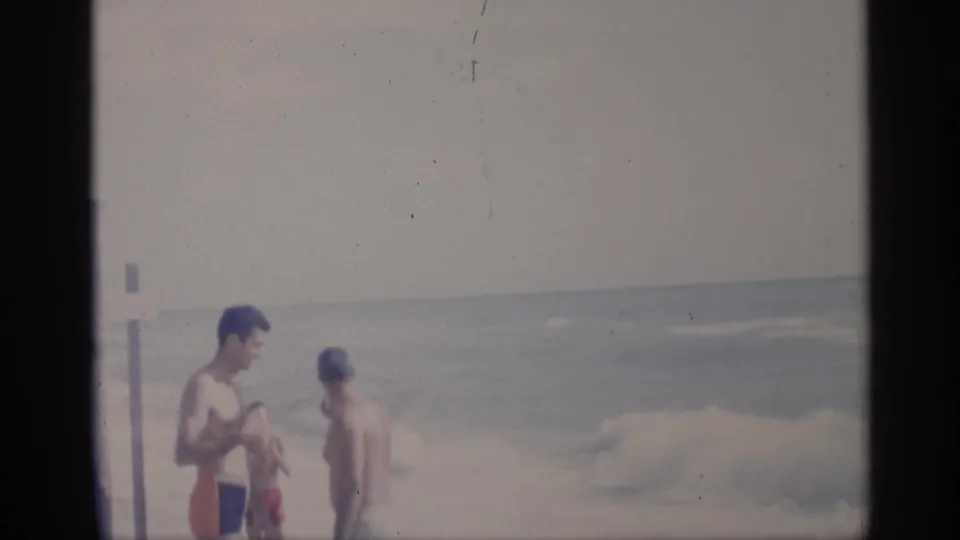 Two adult men pause near the California surf, with a small blurred passerby moving between them and the camera.