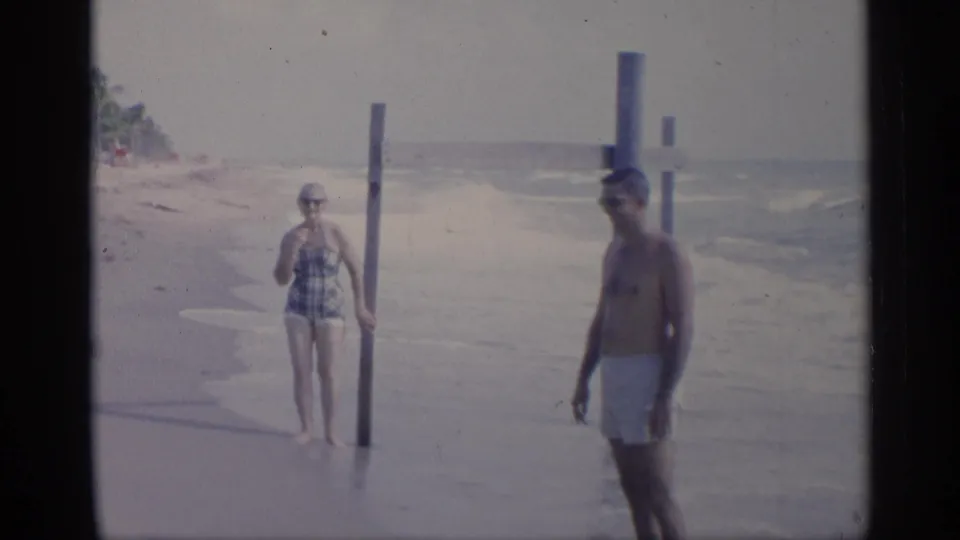 A woman in a patterned one-piece swimsuit stands near a wooden post on a California beach while a man in white trunks walks past.