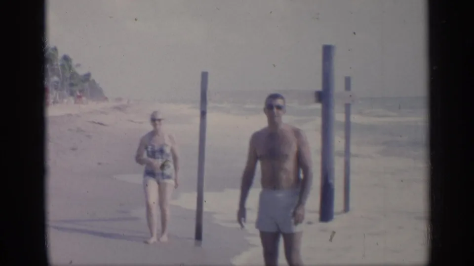 A man in white swim trunks stands in the foreground on a California shoreline, with a woman and upright posts behind him near the surf.