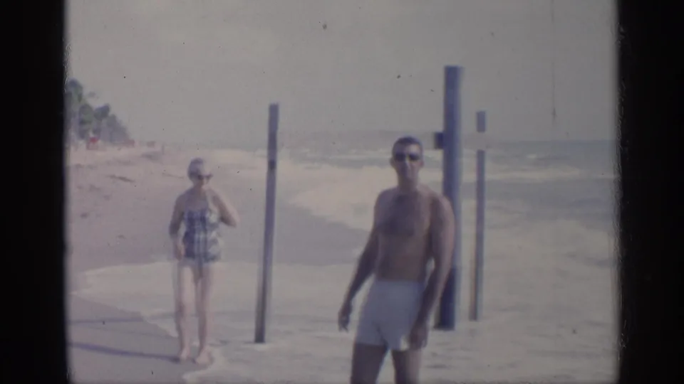 1959 California beach frame with a man in white swim trunks walking toward camera and a woman in plaid swimsuit near rolling surf.