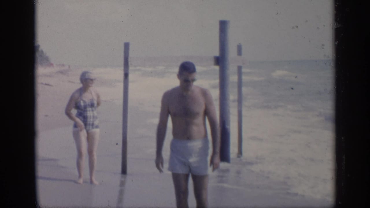 Man in swim trunks walks near the water while a woman stands by beach posts.