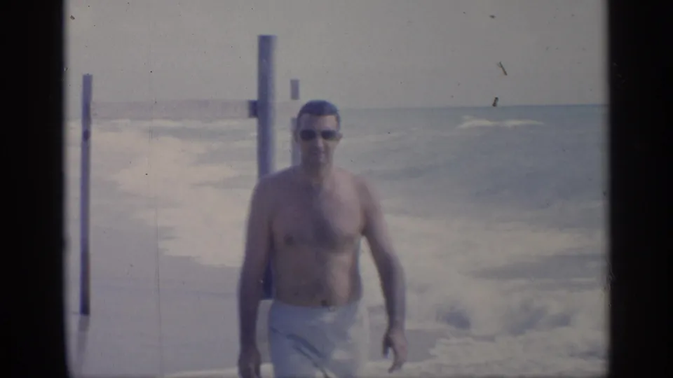 Close 1959 California beach frame of a man in sunglasses and white swim trunks walking in front of churning surf.