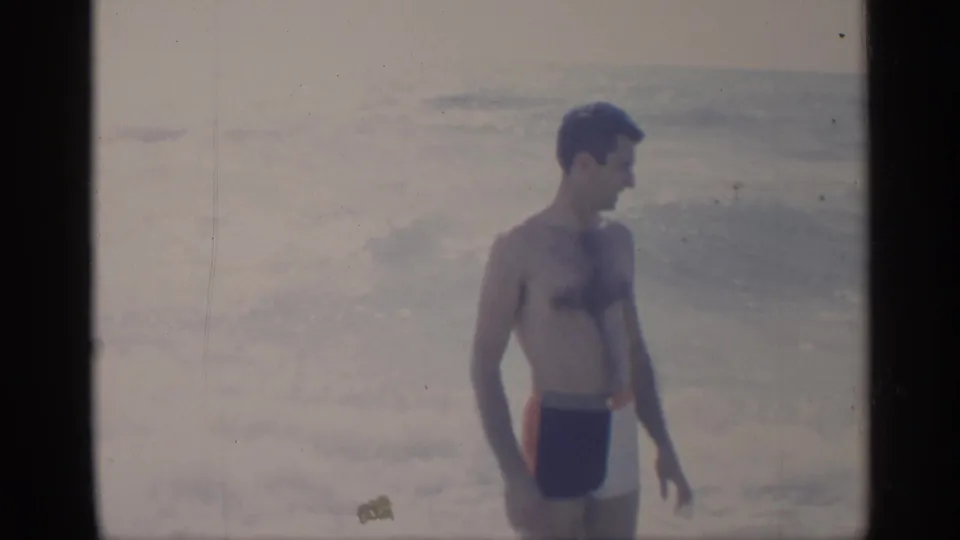 1959 California beach frame showing a man in profile wearing dark swim trunks with red side panels before rough surf.