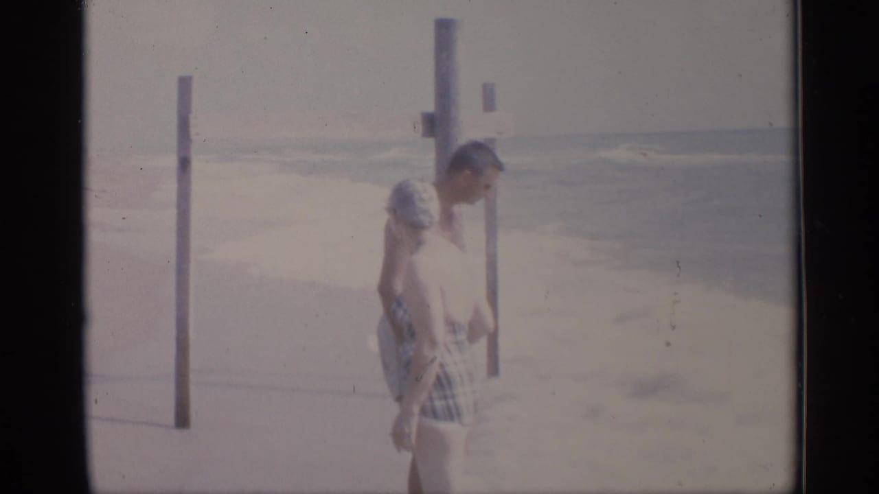 Man and woman in swimsuits walking on sand near posts with ocean surf behind them.