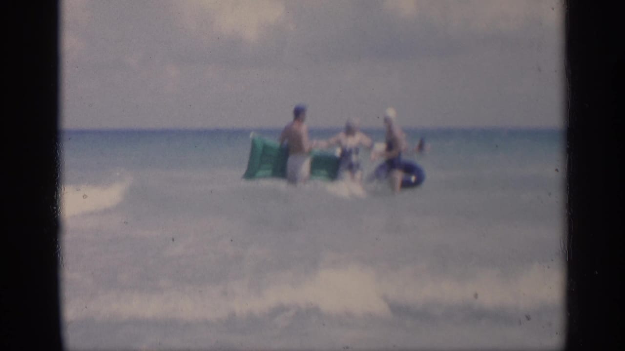 Three adults stand in shallow ocean water holding a green inflatable raft and a blue inner tube.