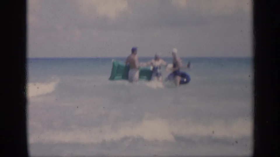 1959 California home-movie frame of three adults in swimwear standing in shallow surf with a green raft and blue inner tube.