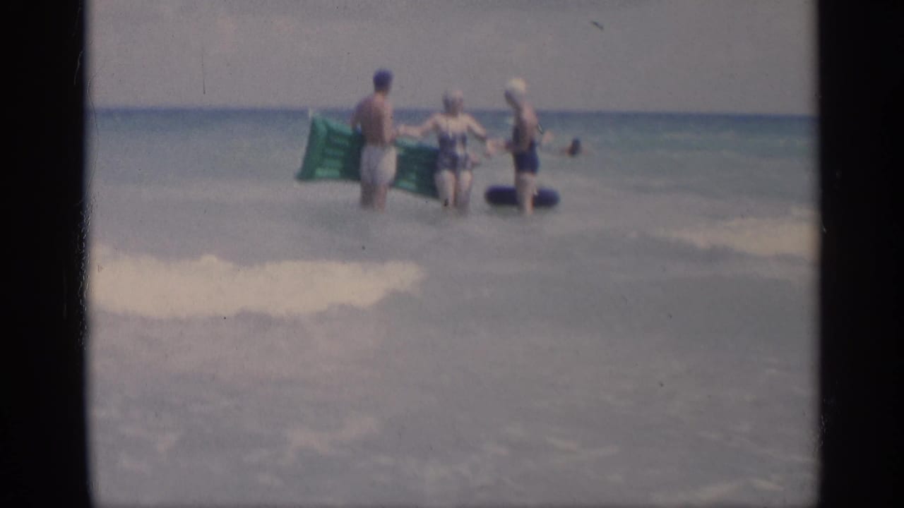 Three adults in swimwear stand in ocean surf with inflatable floats as waves roll behind them.