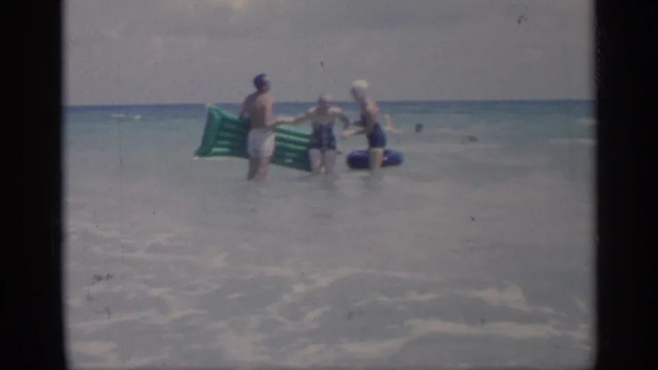 1959 California beach footage frame with adults holding flotation gear in shallow surf and distant swimmers offshore.
