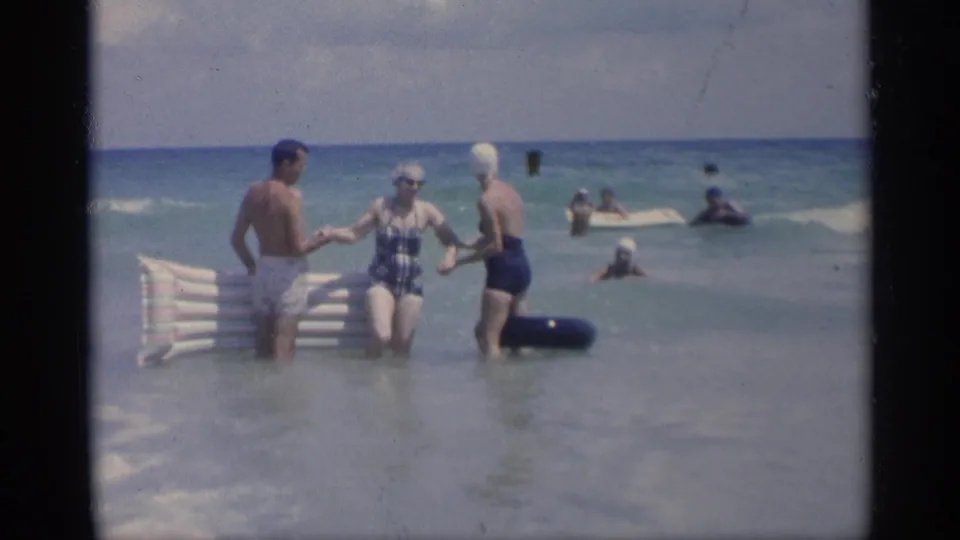 1959 California home-movie frame of three adults in the surf with inflatable beach gear and swimmers in the background.