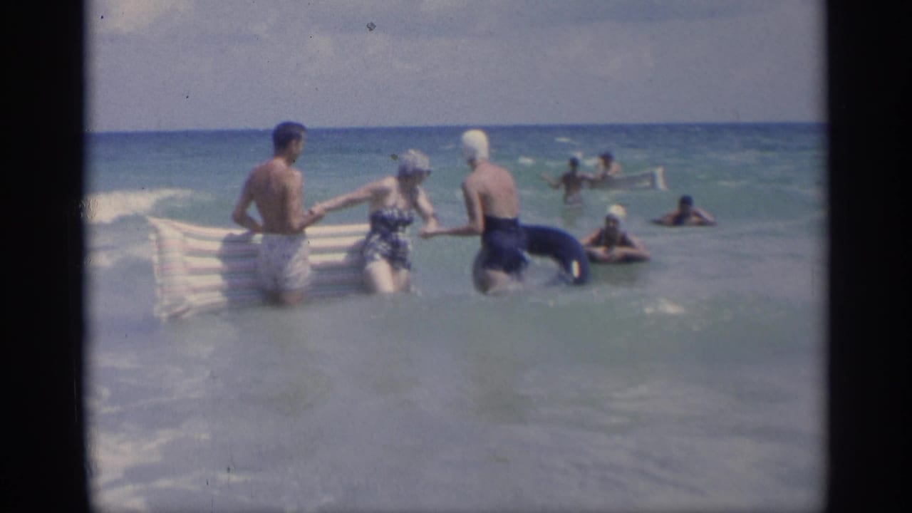 Three adults stand in shallow surf with an inflatable raft and inner tube as swimmers appear offshore.