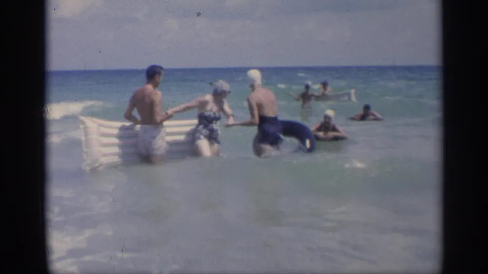 In 1959 California beach footage, three adults hold hands in the shallow surf with a striped raft and dark inner tube nearby.