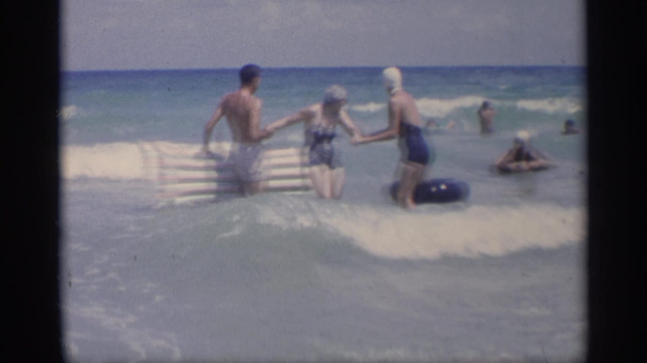 Three adults face the waves while holding hands near a striped raft and dark inner tube.
