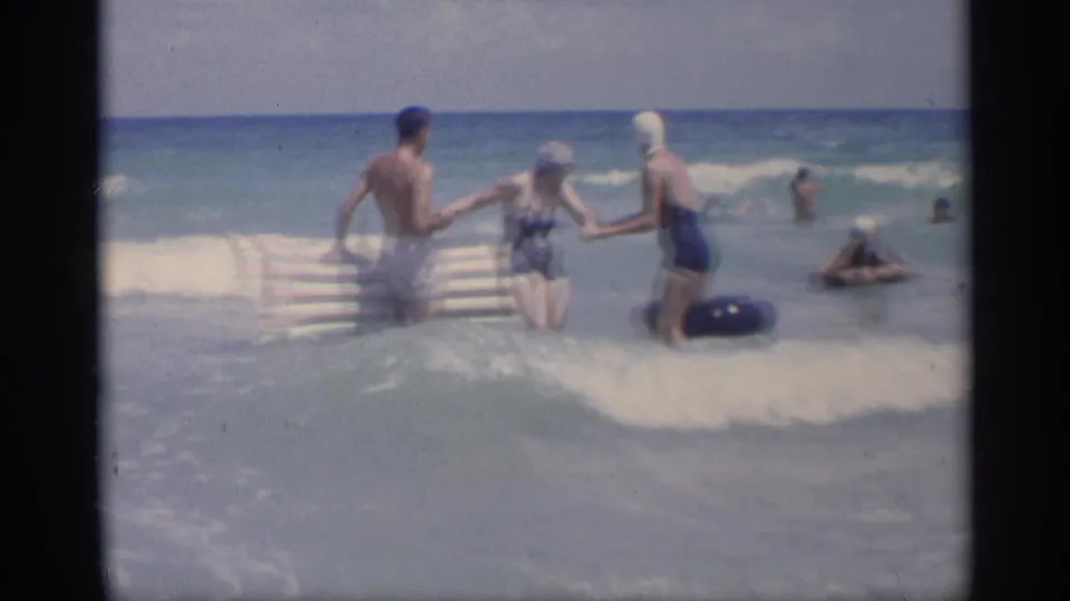 Three adults in mid-century swimwear stand in California surf, with a striped inflatable raft and blue inner tube beside them.