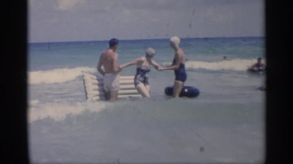 The beachgoers remain linked in shallow California surf while a wave curls behind the inflatable raft and inner tube.