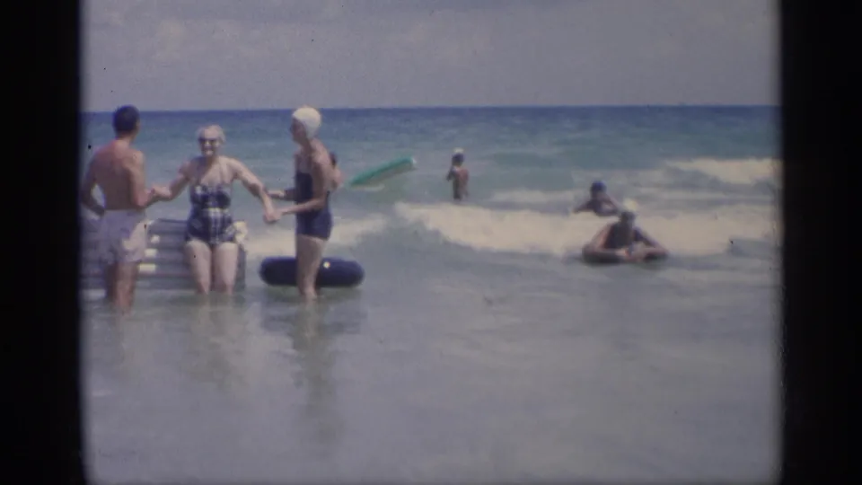 A woman in a patterned swimsuit and swim cap steps forward in the 1959 California surf, flanked by two companions and beach inflatables.