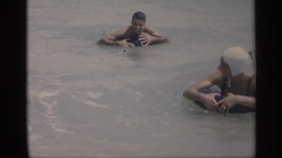 1959 California beach footage shows swimmers resting in the water, with a white swim cap visible at the edge of the frame.