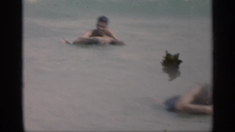 1959 California 8mm frame with a swimmer in shallow surf and seaweed floating in the foreground water.