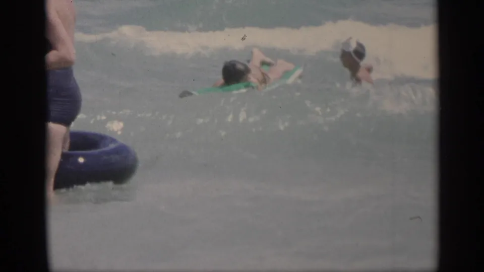 1959 California ocean scene with swimmers and flotation rings near a breaking wave, captured in soft handheld 8mm color.