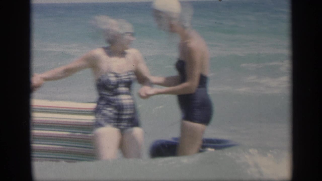 Two women in swimsuits hold hands in shallow ocean surf beside a striped beach mat.
