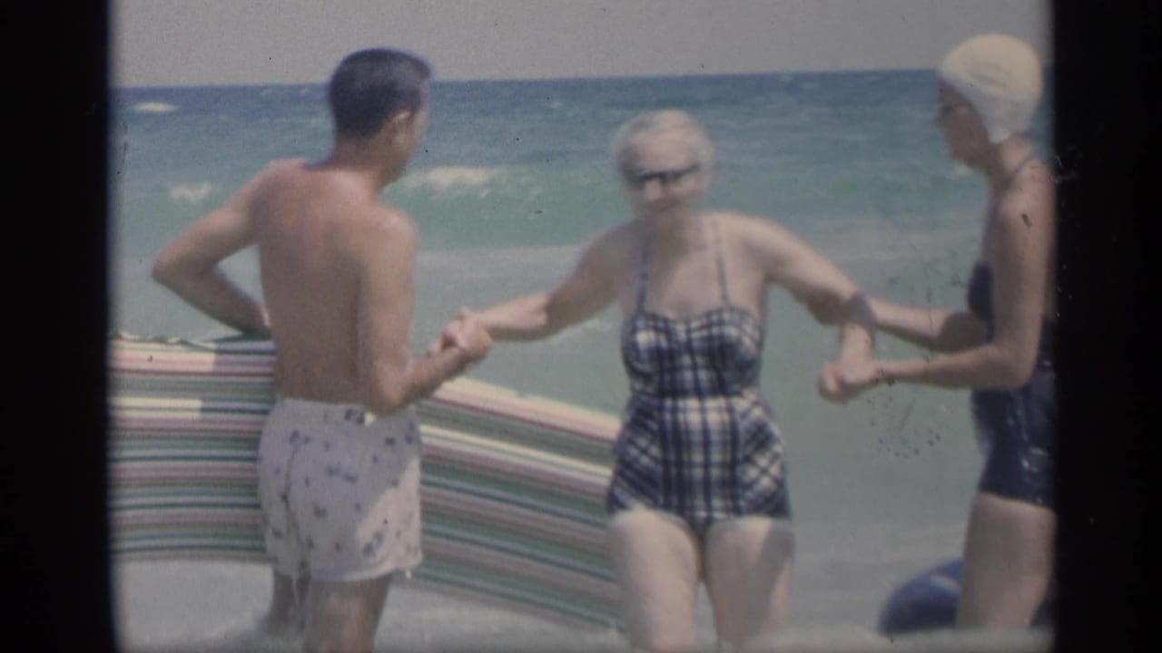 A man and two women stand in ocean waves, holding hands near a striped beach mat.