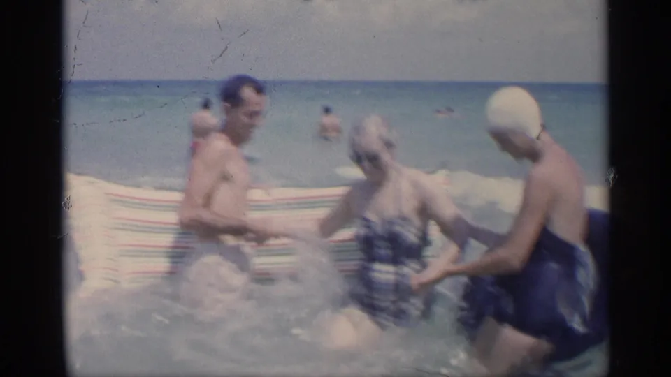 1959 California beach frame showing three adults laughing or moving in shallow waves, with distant swimmers and open water behind them.