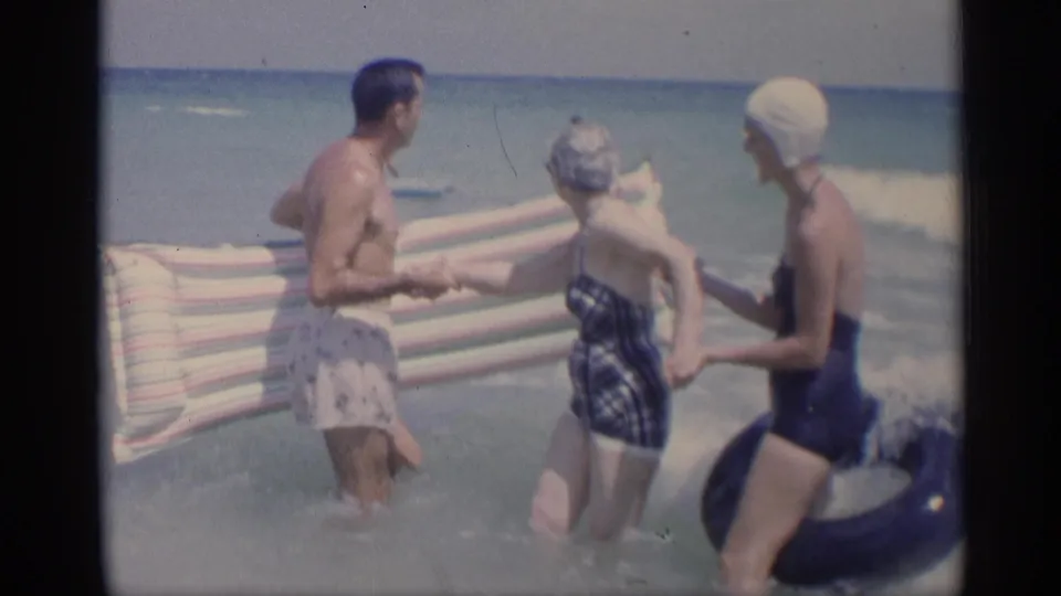 1959 California beach home movie frame with three adults holding hands in the surf, one woman beside a dark inner tube.
