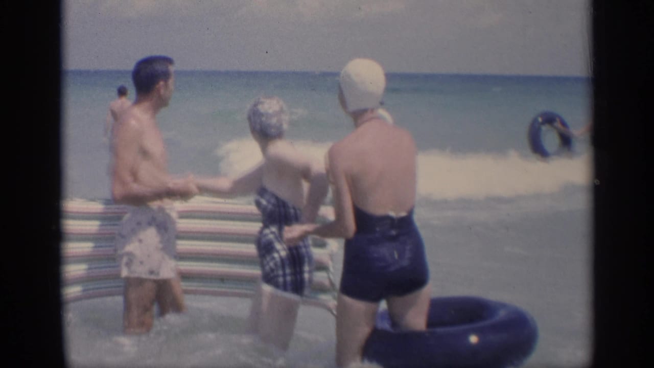 Three adults wade in ocean surf with a striped beach mat and inflatable ring.