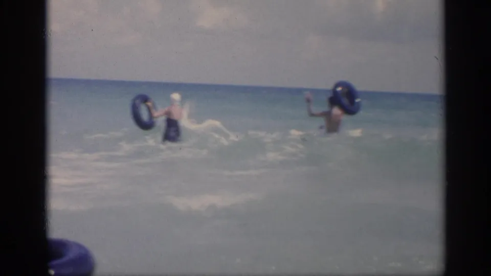 Two adults appear farther out in the California surf, each near a blue inflatable ring as waves break around them.