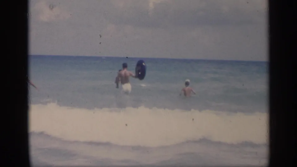 California beach home-movie frame with adults wading into the ocean while a white wave breaks across the foreground.
