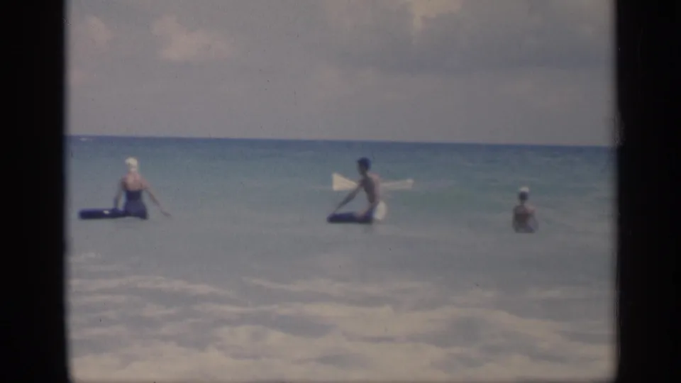 Three adults are visible in shallow California surf, with blue inflatable rings near two of them and white foam in the foreground.