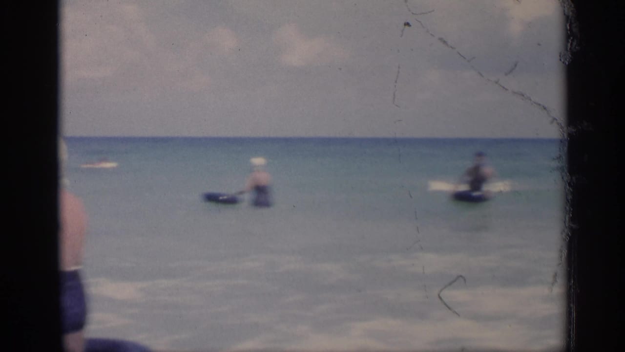 Distant swimmers and beachgoers in shallow ocean water with film gate borders visible.