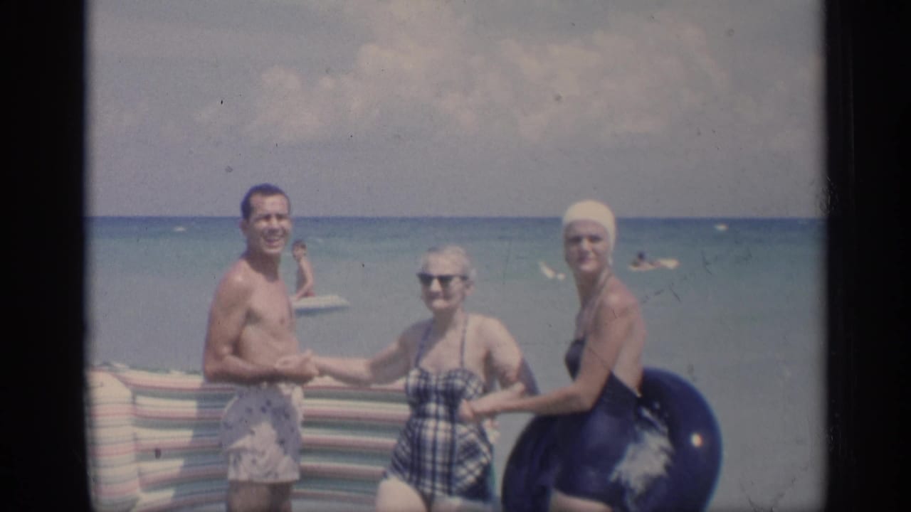 Three adults in swimsuits stand in shallow surf beside a striped beach blanket and swim tube.