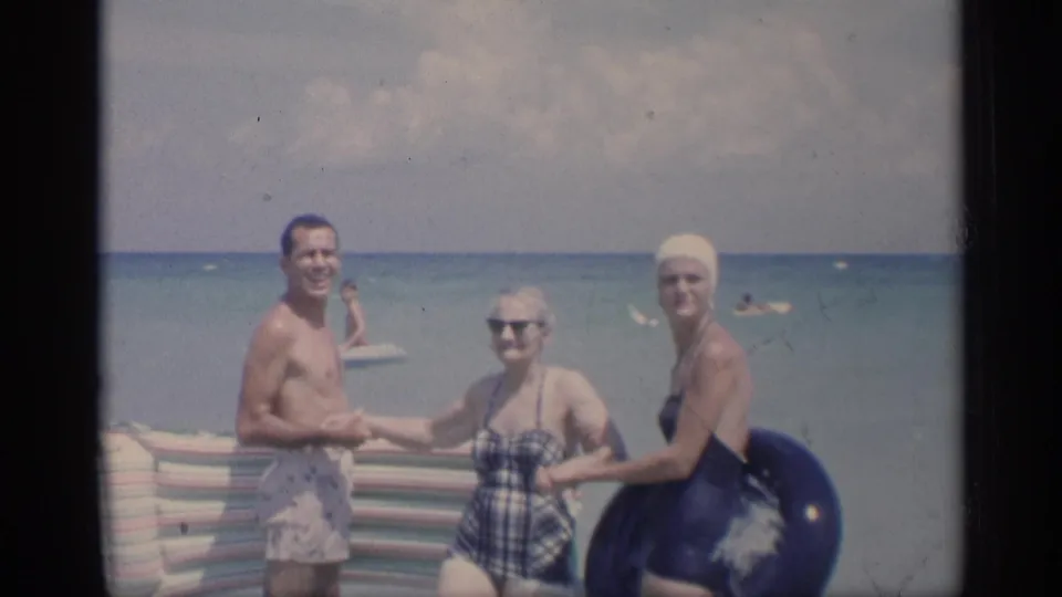 1959 California beach frame with three adults in period swimsuits posing in shallow surf beside a striped blanket and dark swim tube.