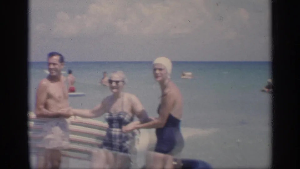 1959 California beach home-movie frame showing three adults smiling near a striped blanket, with ocean water and swimmers behind them.