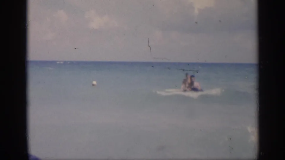 1959 California beach frame showing two people floating offshore on an inflatable, with calm water, horizon, and sky in view.