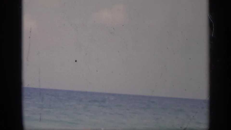 1959 California beach footage frame with the ocean horizon, muted blue water, pale sky, and visible 8mm contact-sheet borders.