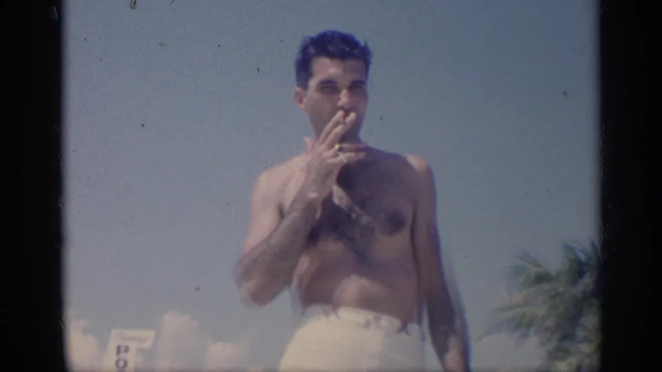 1959 California home-movie frame showing a shirtless adult man in light swim trunks smoking near the beach, with sky and palm foliage behind him.